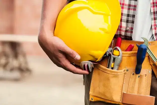 A construction worker holding his yellow hard hat next to a work belt full of tools. 