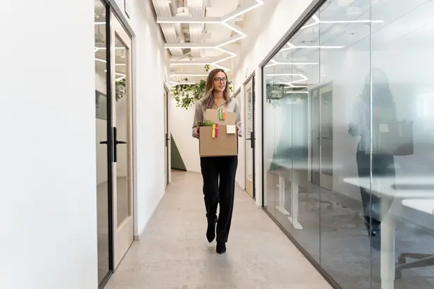 A white business woman carrying a box of supplies through an office building. 