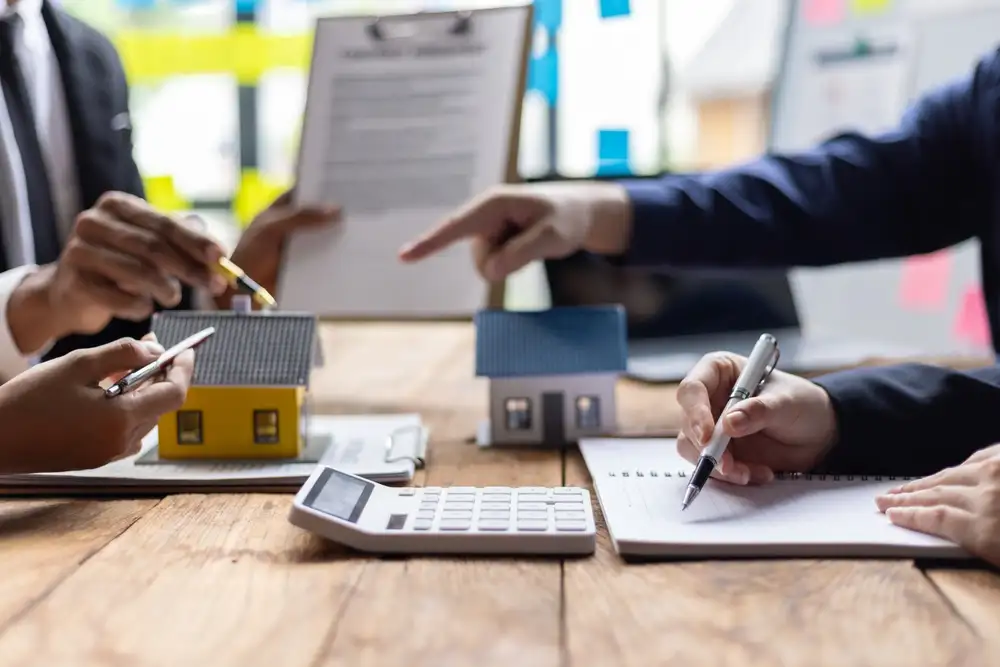 Real estate agent calculates property value with customer, discussing investment potential surrounded by paperwork. Consultation on buying, selling, or renting.