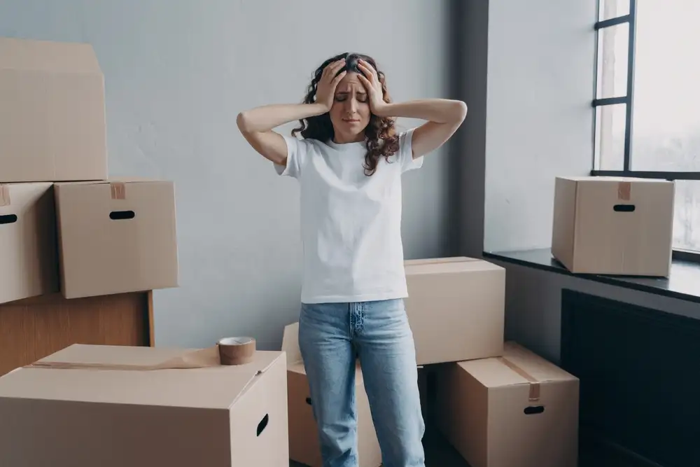 Headache from moving. Exhausted woman is tired from packing cardboard boxes with duct tape. European young lady in jeans and white t-shirt going to relocate. Anxiety and stress from moving concept.